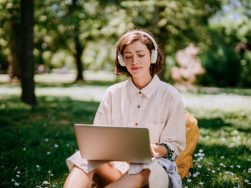 Eine Frau sitzt mit Kopfhörern und Laptop auf einer Wiese.